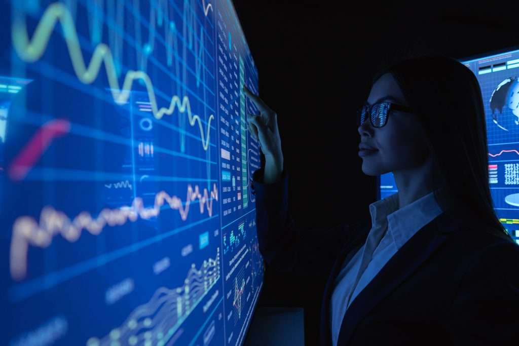 Person in a dark room examining a wall of blue stock charts and graphs, pointing at data displays with a focused expression